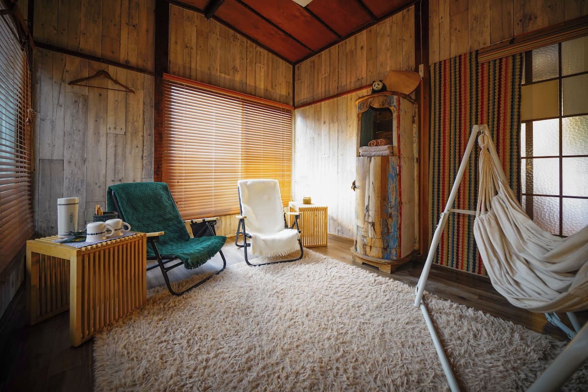 Una habitación con paredes de madera, una hamaca, una silla verde y una silla blanca, además de un mueble pequeño.