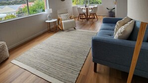 A modern living room with a large window, a blue sofa, a dining table, and a view of a body of water.