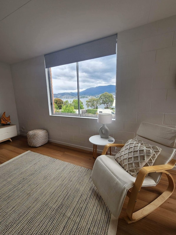 Una habitación moderna con un ventanal grande, una mecedora, una mesita y un sofá blanco.
