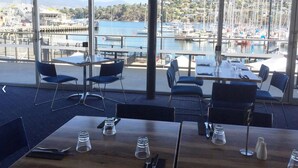 A restaurant with large windows overlooking a marina with boats. Tables and chairs are arranged for dining.