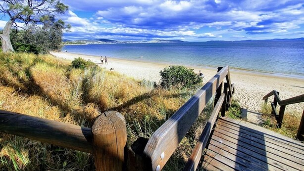 A wooden boardwalk leading to a beach with a clear view of the sea and distant mountains.