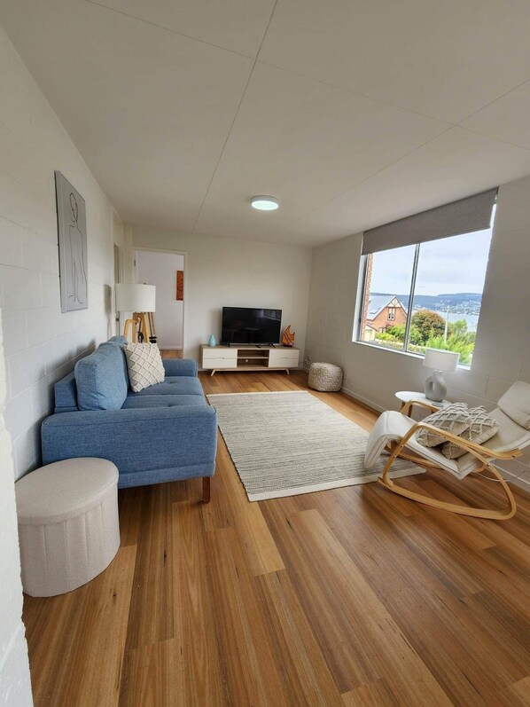 Una sala moderna con un sofá azul, un sillón blanco, piso de madera, televisor y un ventanal con vistas.