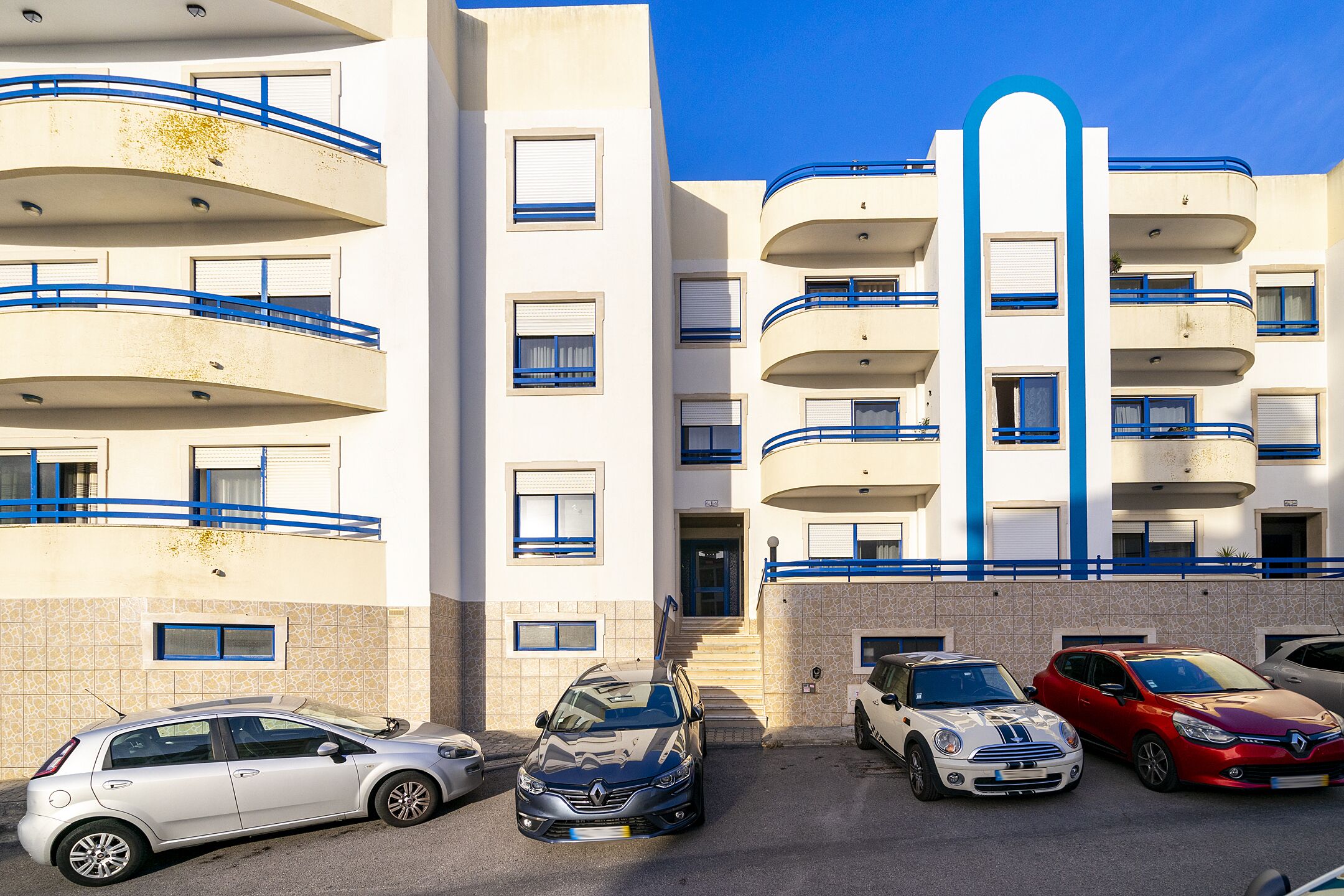 A multi-story residential building with balconies, a central stairwell, and several parked cars.