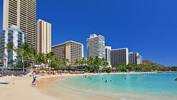 A beach with clear turquoise waters, palm trees, and tall buildings in the background. - Great Partial Ocean View! Full Kitchen, Free Parking, Pool! Heart of Waikiki! (Honolulu)