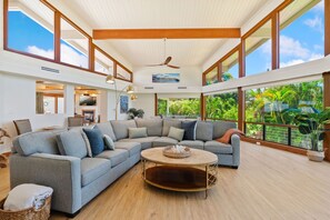 Un salon spacieux avec un grand canapé d’angle, une table basse ronde et un ventilateur de plafond. De grandes fenêtres offrent une vue sur la végétation luxuriante à l’extérieur.