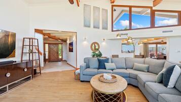 A spacious living room with a large sectional sofa, a round coffee table, and a TV on a wooden cabinet. There are multiple light fixtures and a large window providing natural light.