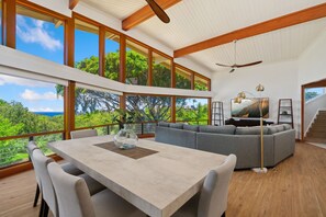 Un vaste espace de vie avec une grande fenêtre donnant sur un paysage luxuriant, une table à manger avec des chaises et un canapé d’angle gris.