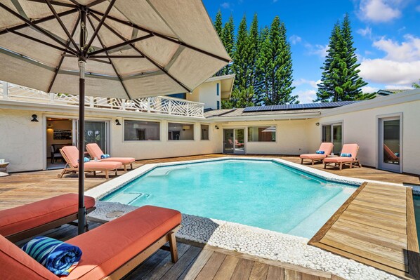 A swimming pool with lounge chairs and an umbrella, surrounded by a wooden deck and white pebbles.