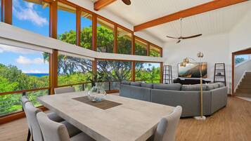A spacious living area with a large window overlooking a lush landscape, a dining table with chairs, and a gray sectional sofa.