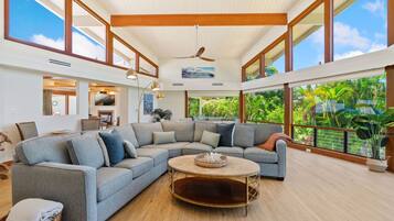A spacious living room with a large sectional sofa, a round coffee table, and a ceiling fan. Large windows offer views of lush greenery outside.
