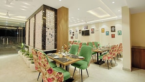 A restaurant with green upholstered chairs, a wooden table, and a decorative wall with framed pictures. - Veda Inn By Casa Ekam (Yamkeshwar)