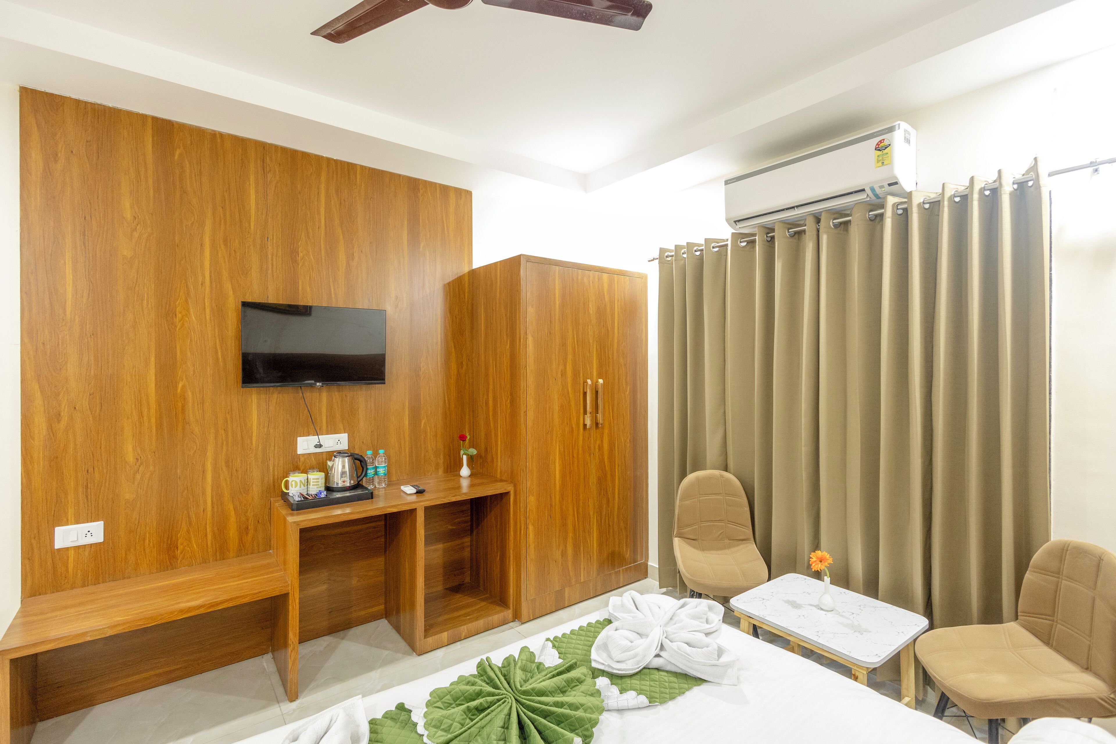 Hotelkamer met een houten garderobe, een aan de muur bevestigde televisie, een bureau met stoel, een bed met een bladmotief en een klein tafeltje met een bloevas.