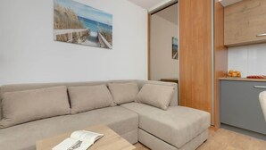 A modern living room with a beige sofa, a wooden coffee table with a book, a painting of a beach scene, and a kitchen area with a grey cabinet and wooden countertop.