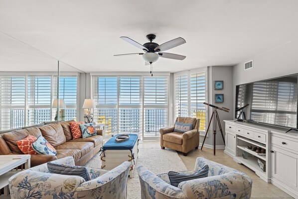 A spacious living room with a large window, a ceiling fan, a television, and a white cabinet. - Recently updated oceanfront unit, wonderful views + Free Attraction Tickets! (North Myrtle Beach)
