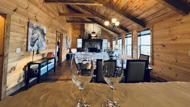 Interior de una cabaña de madera con comedor que incluye una mesa, dos copas de vino, chimenea de piedra y paredes de madera.
