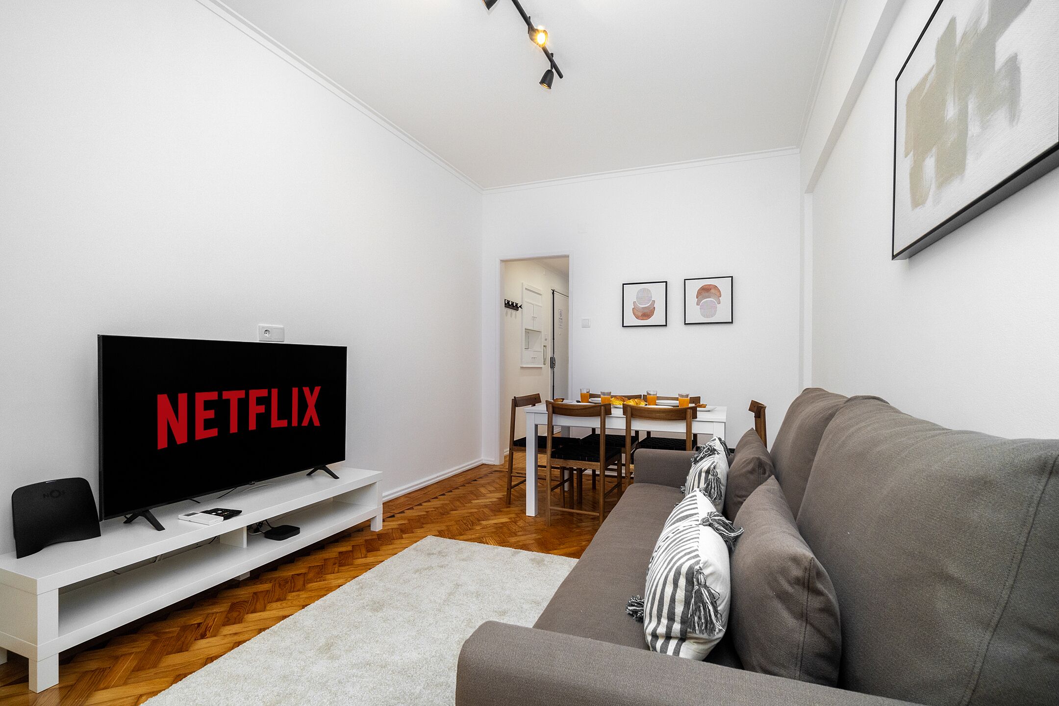 A modern living room with a flat-screen TV, a grey sofa, a white entertainment unit, and a dining area with a wooden table and chairs.