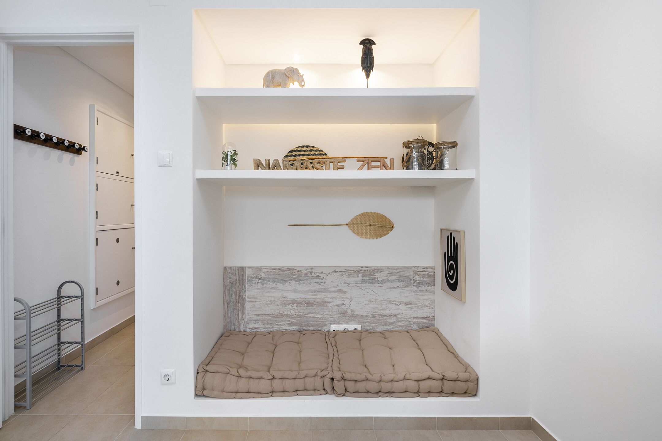 Une pièce avec un mur blanc, une étagère ornée d’objets décoratifs, un luminaire fixé au mur et une autre étagère sur laquelle se trouve un panier en osier.