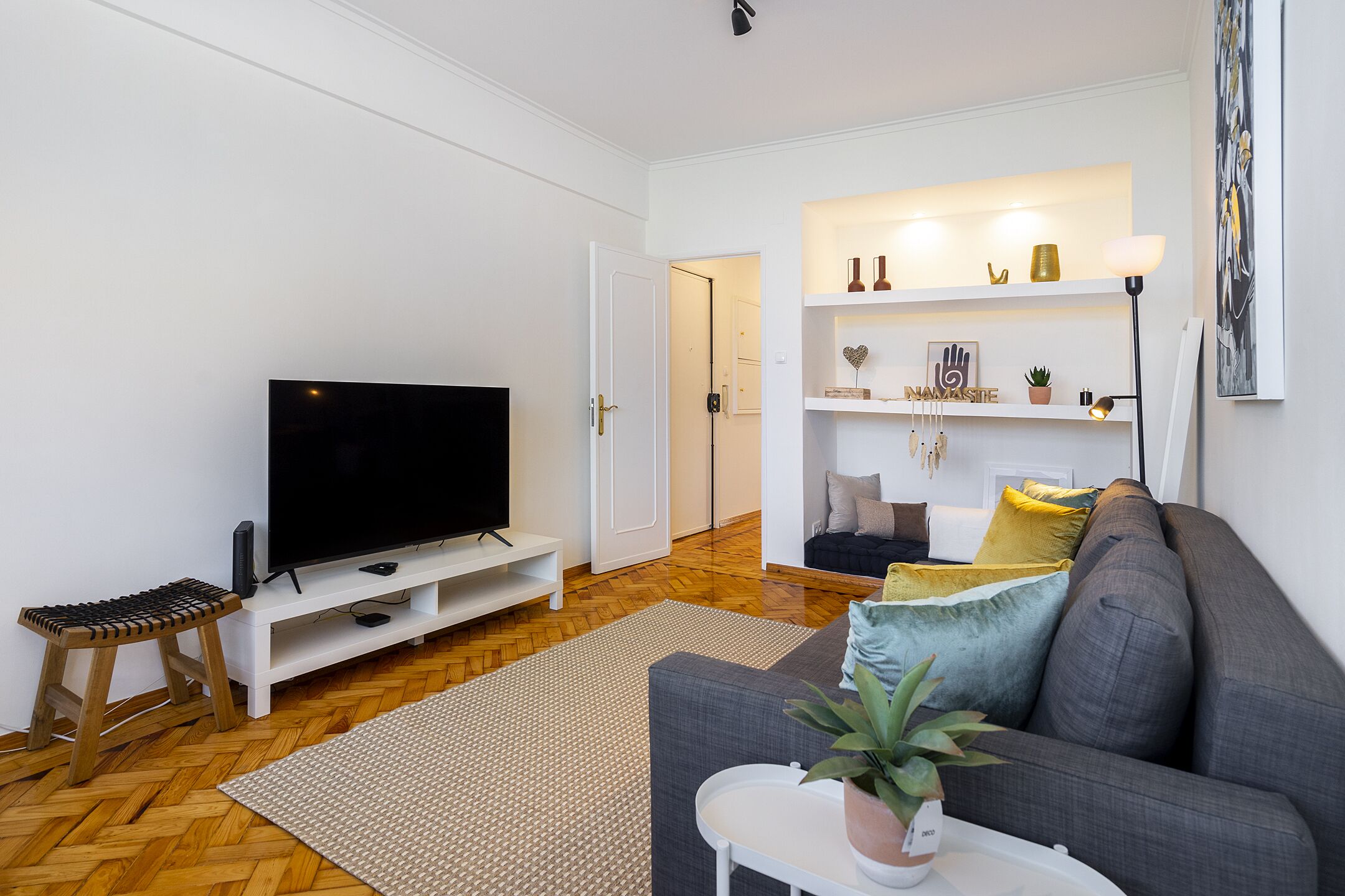 A modern living room with a flat-screen TV, a grey sofa, a small white table with a plant, and a wooden stool.
