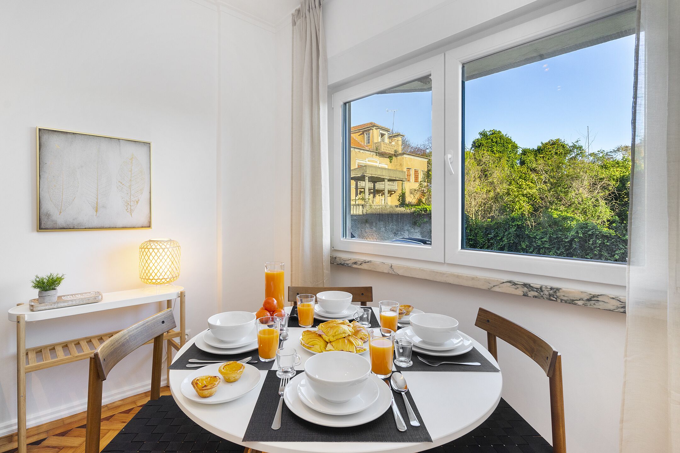 A breakfast table set with white plates, bowls, and cutlery, a lamp, and a view of greenery and buildings through a window.