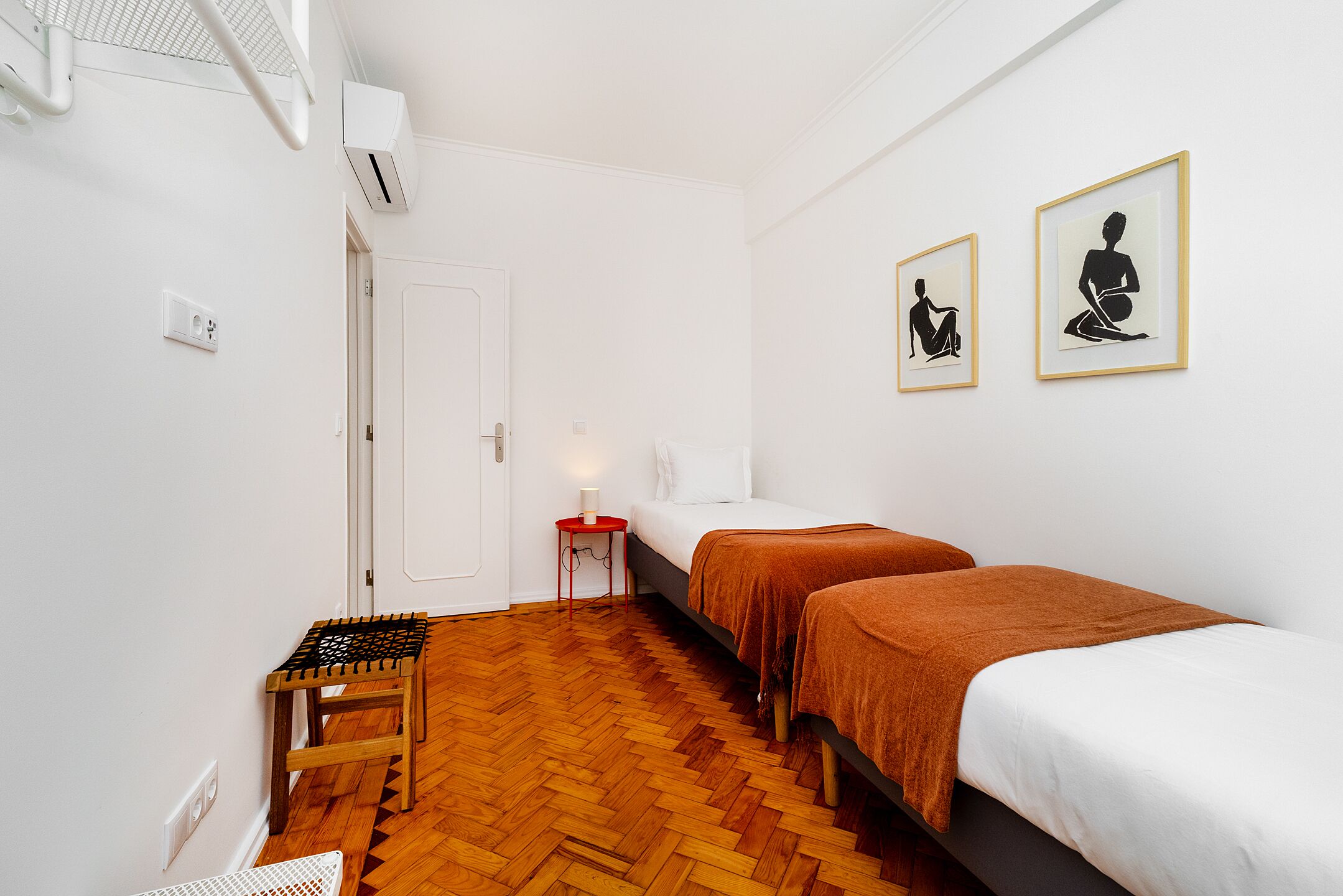 Une pièce avec deux lits, un banc en bois, une petite table avec une lampe et deux tableaux encadrés accrochés au mur.