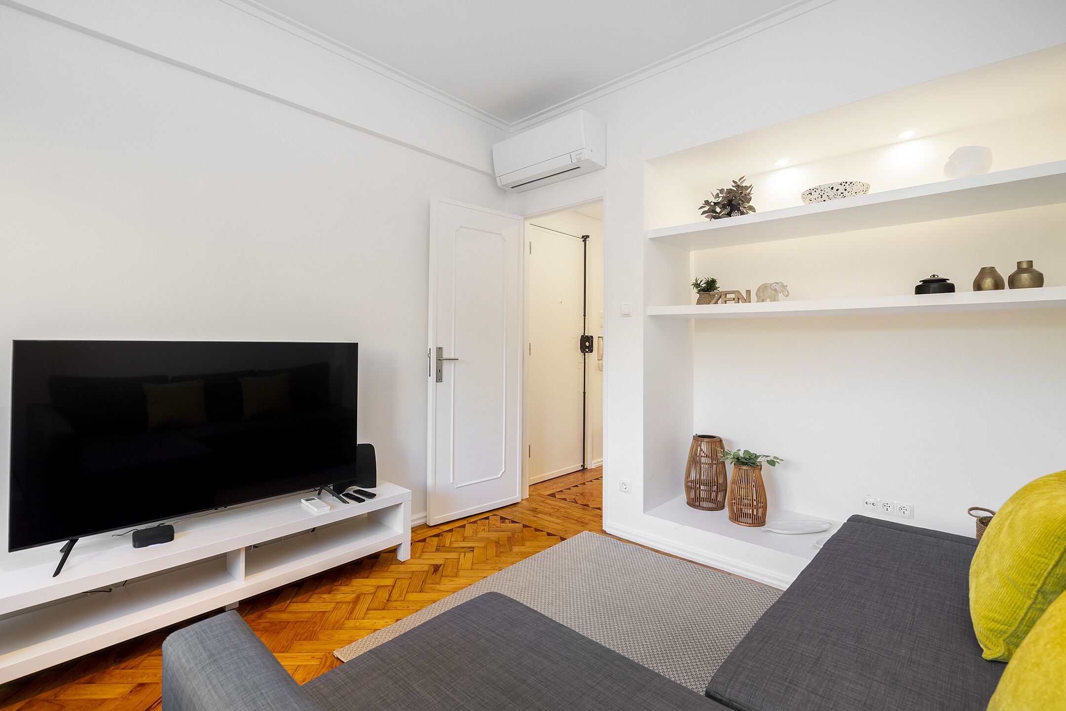 Un salon moderne avec un téléviseur à écran plat posé sur un meuble blanc, un canapé gris et une petite table sur laquelle se trouve une télécommande.