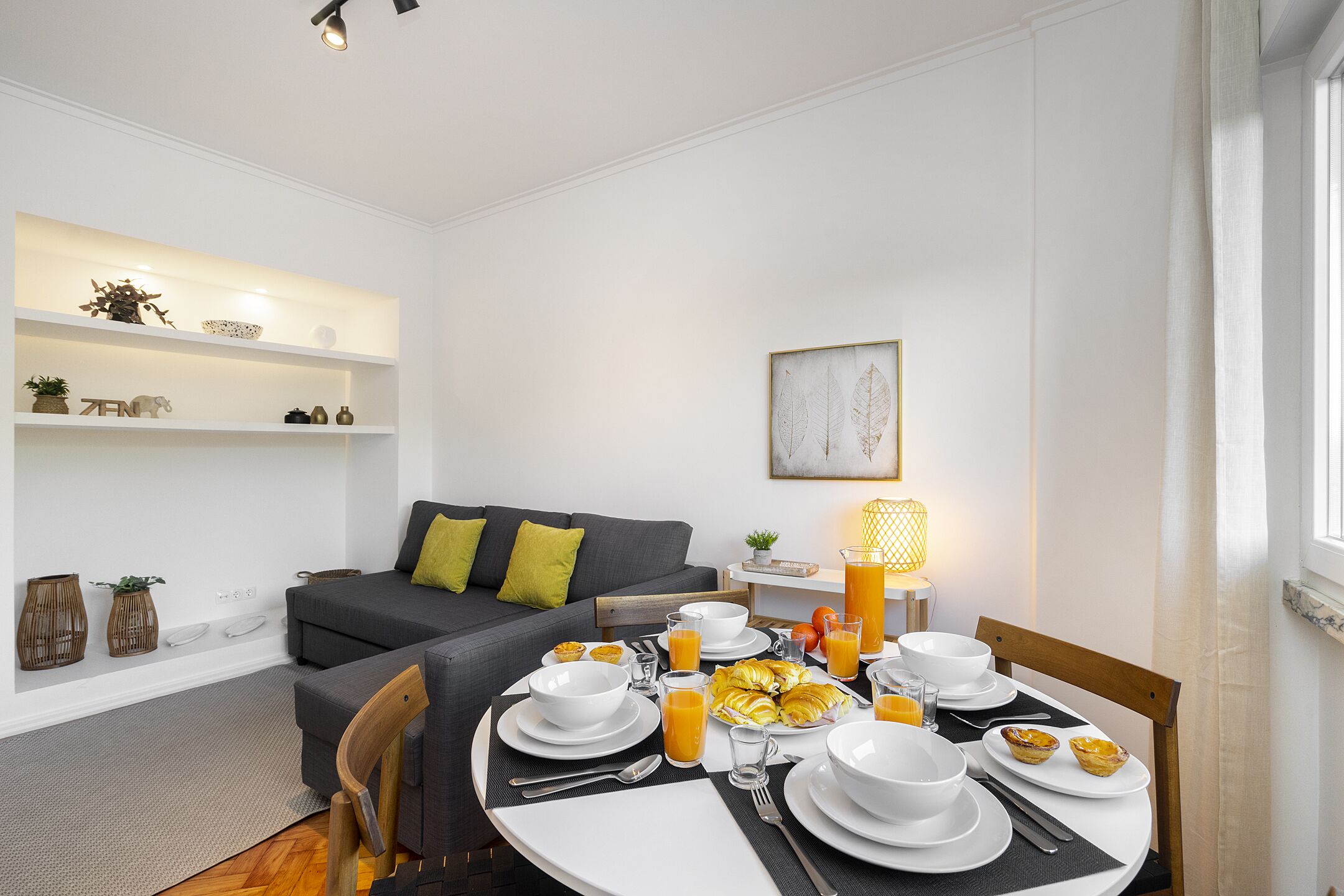 A modern living room with a dining area, featuring a sofa, a dining table set with plates, glasses, and a lamp.