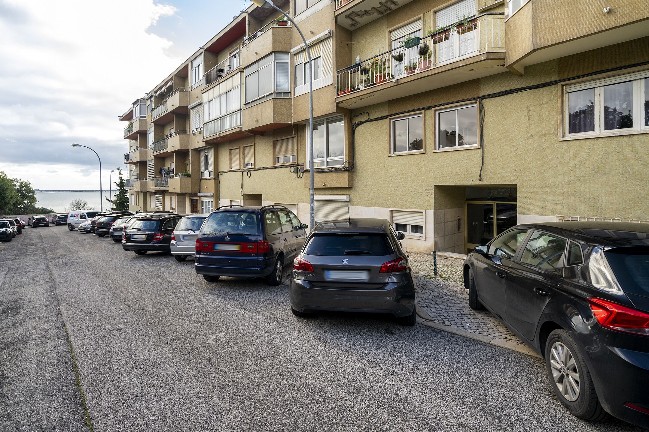 A row of parked cars along a street with a multi-story building featuring balconies with plants.