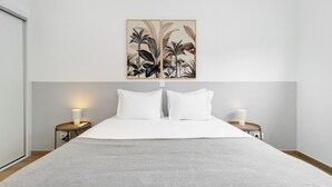 A neatly made bed with white linens, two bedside tables with lamps, and a wall-mounted artwork of tropical plants. - Peaceful Algés Family Home (Oeiras)