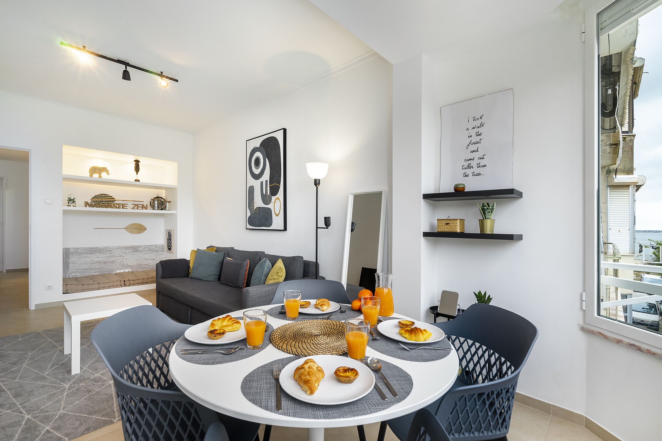 A modern living room with a dining area, featuring a sofa, a coffee table, and a dining table set for breakfast with croissants and orange juice.