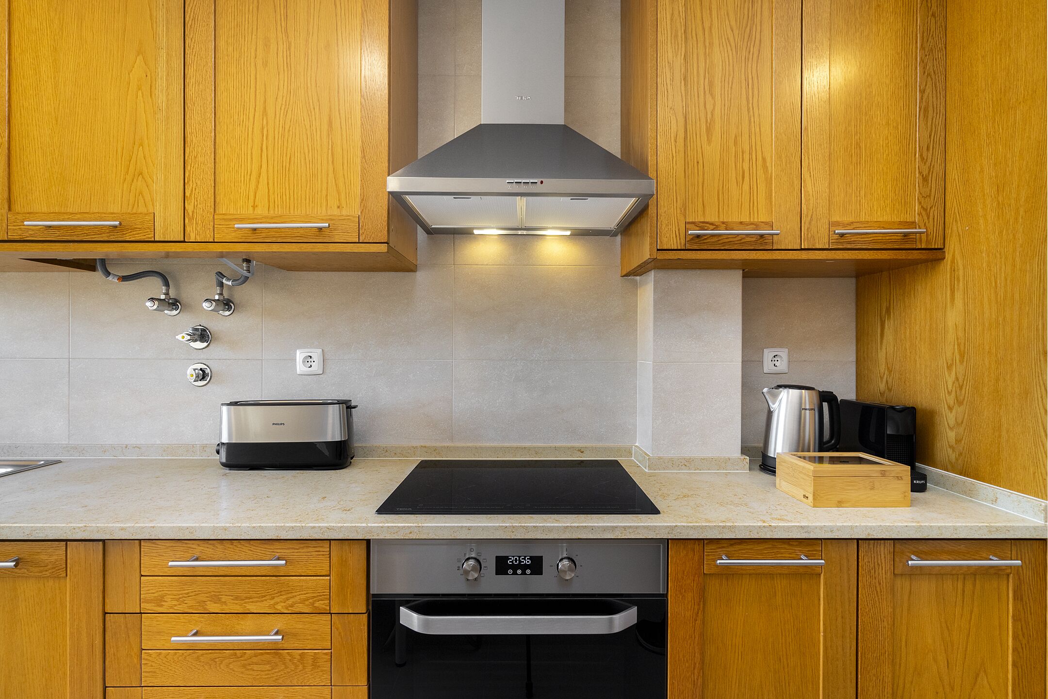 A modern kitchen with wooden cabinets, a stainless steel range hood, a built-in oven, and a black induction cooktop.
