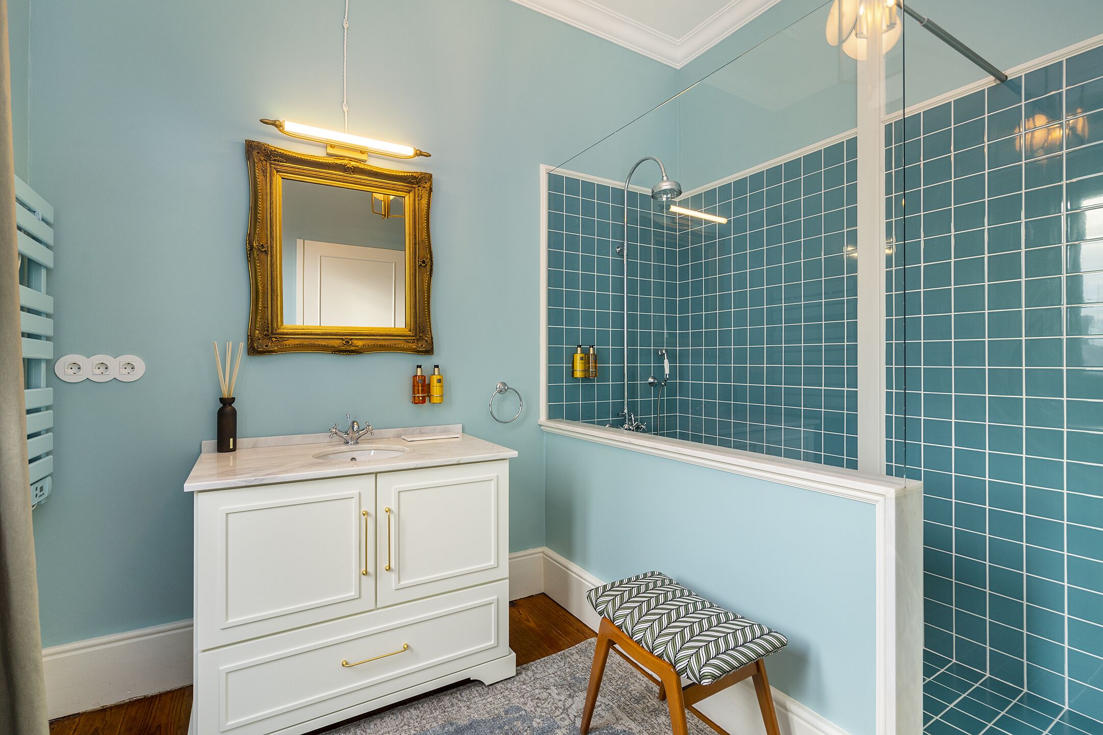 Un baño con tocador blanco, un espejo con marco dorado y ducha con azulejos azules.