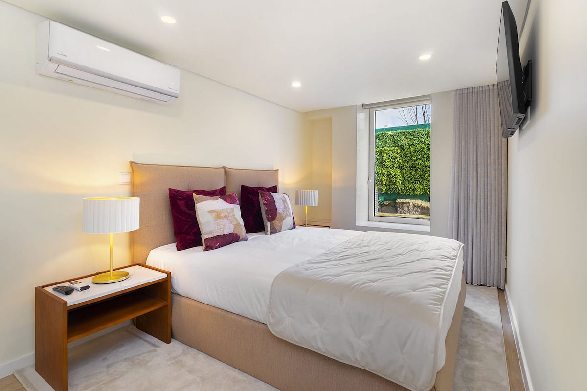A modern bedroom with a large bed, bedside lamps, a nightstand, and an air conditioning unit.