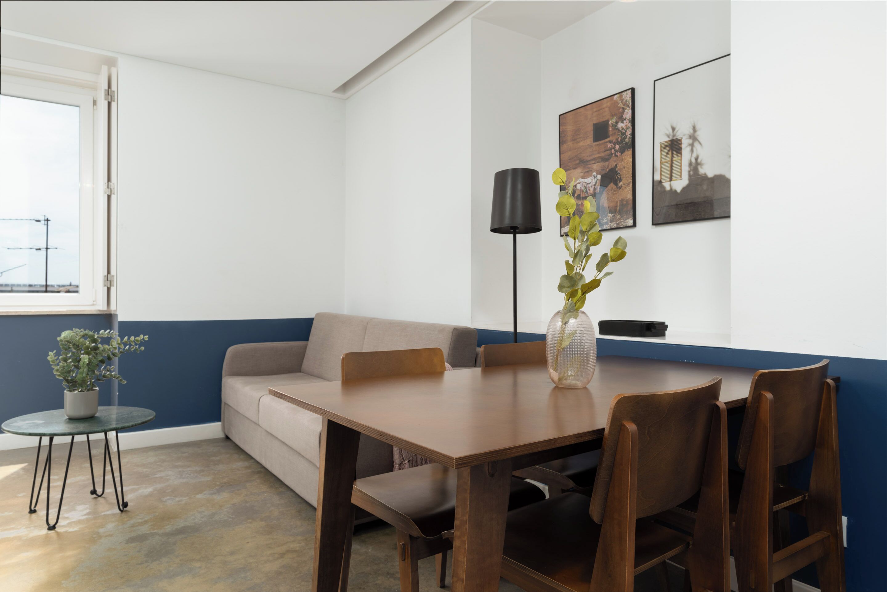 Un salon moderne avec une table à manger en bois, deux chaises, un canapé beige, une petite table ronde ornée d’un pot de plantes et un lampadaire.