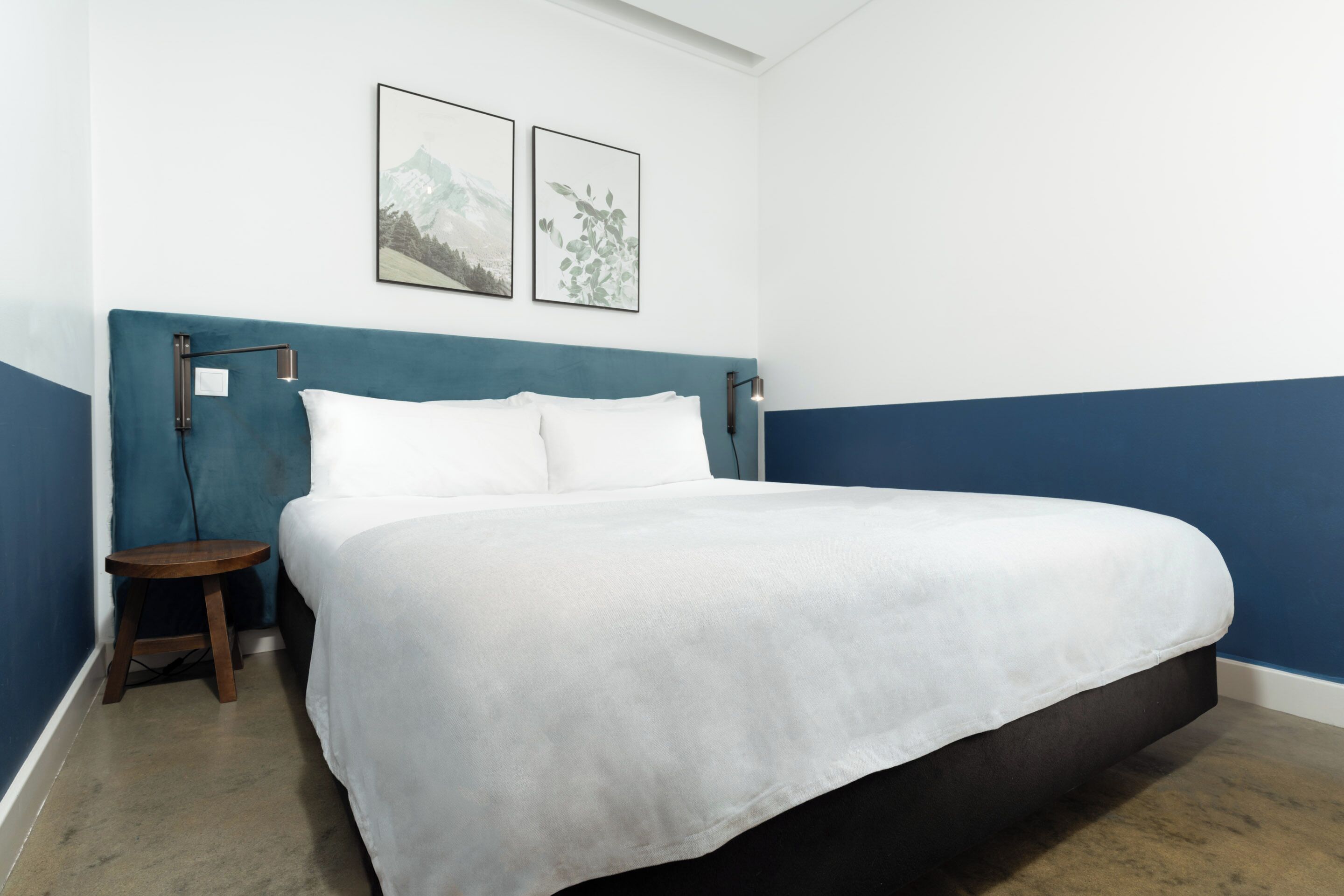Una cama bien hecha con ropa blanca, una mesita de noche de madera y dos cuadros en la pared.