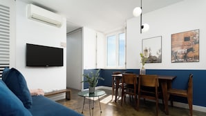 Una sala moderna con un sofá azul, una pequeña mesa redonda con una planta, un área de comedor con sillas de madera y un televisor montado.