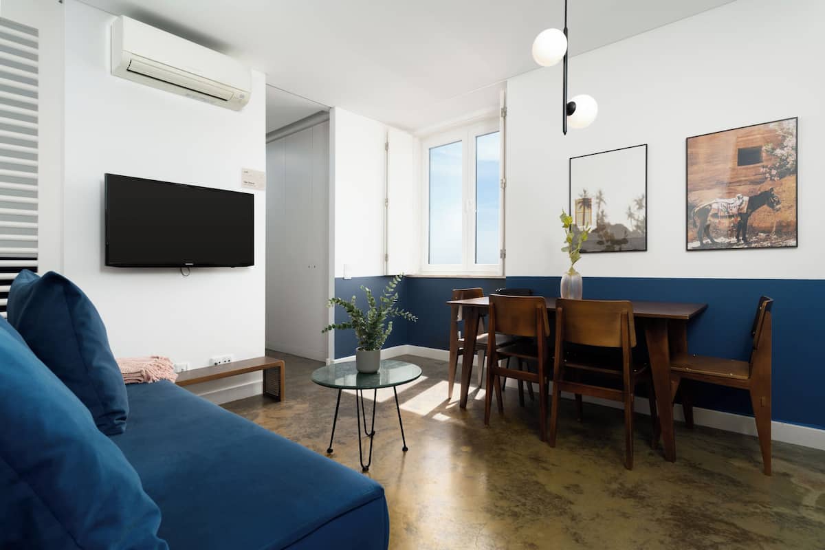 Un salon moderne avec un canapé bleu, une petite table ronde agrémentée d’une plante, un coin repas avec des chaises en bois et un téléviseur fixé au mur.