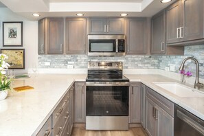 A modern kitchen with stainless steel appliances, marble countertops, and wooden cabinets. - Kamaole Sands 6-101 Completely Renovated Garden View Inner Court Central AC! (Kihei)