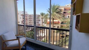 En balkon med en træstol og udsigt over et boligområde med palmer og høje bygninger.