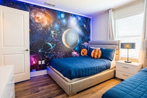 A bedroom with a bed, a nightstand, a wall mural of space, and a window with blinds.