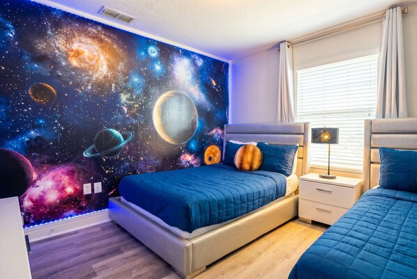 A bedroom with a galaxy-themed wall mural, a bed with blue bedding, a bedside table, and a window with blinds.