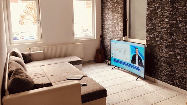 Una sala de estar con un sofá, un televisor mostrando un noticiero, una guitarra y una pared con textura de piedra.