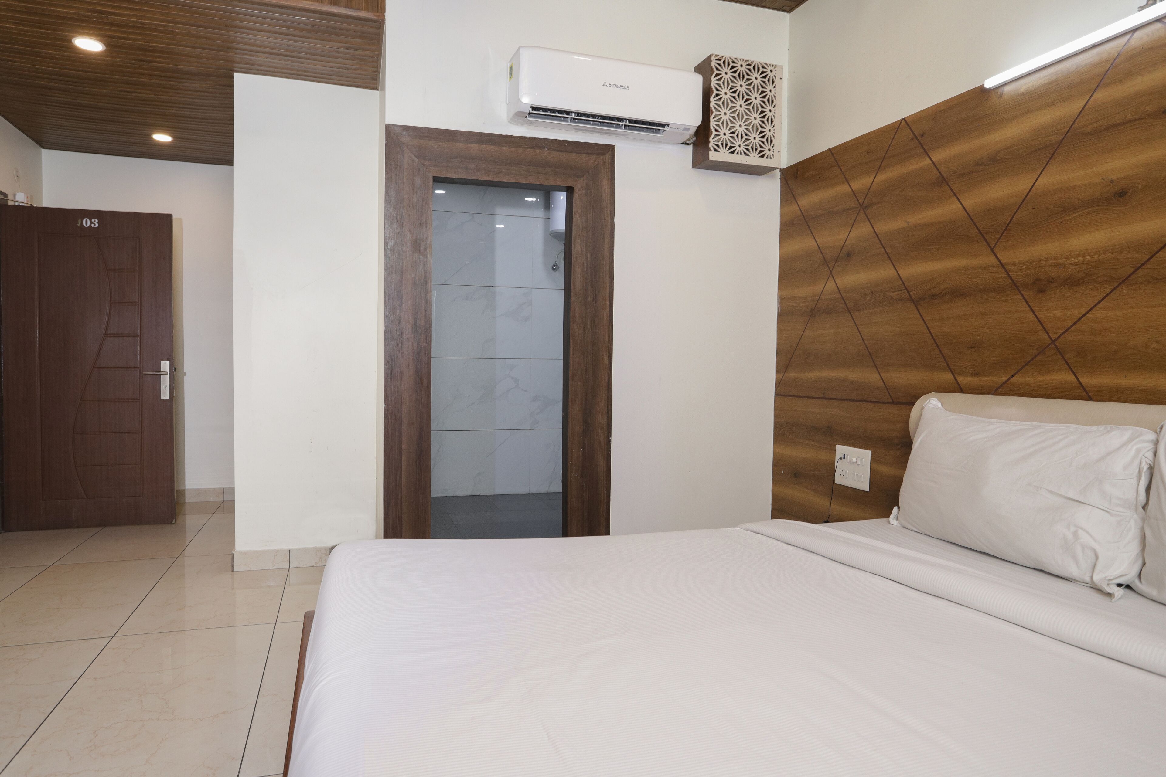 Een hotelkamer met een houten hoofdeinde, een bed met witte lakens, een deur met een nummer en een airconditioning.