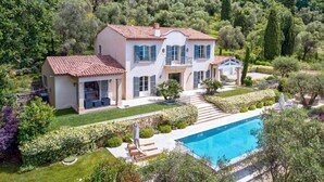 A large, two-story house with a swimming pool, surrounded by lush greenery and hills.