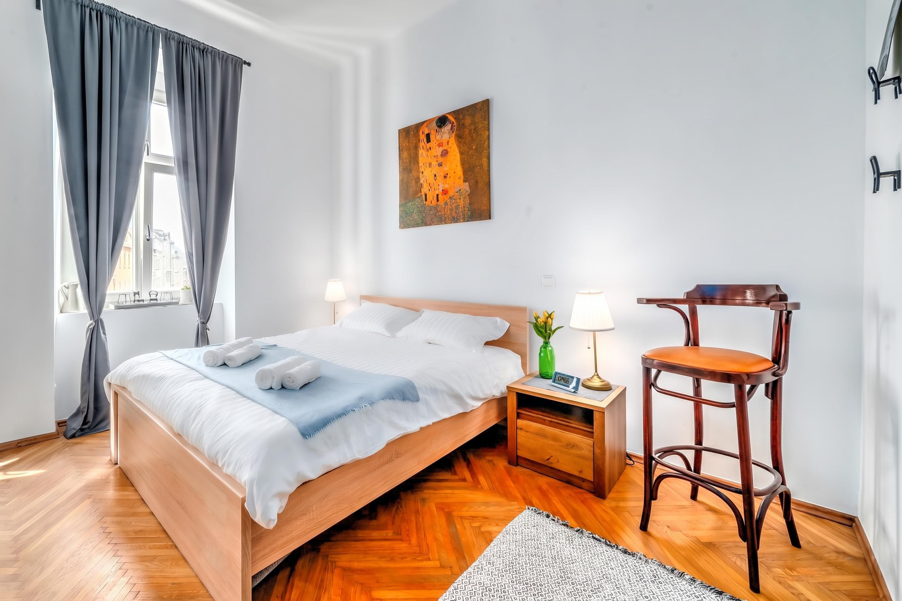 Una habitación de hotel con una cama de madera, una mesita de noche con lámpara, una silla y un cuadro en la pared.
