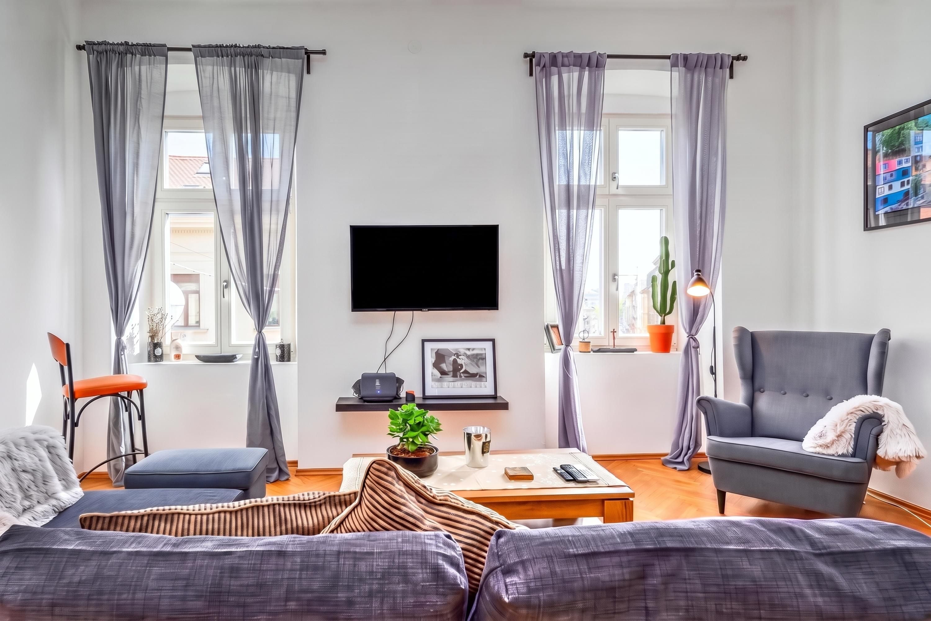Un salon équipé d’un téléviseur à écran plat, de deux canapés, d’une table basse et d’un fauteuil. Il y a deux fenêtres avec des rideaux, une plante en pot et un tableau encadré au mur.