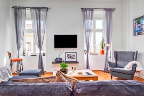 Sala de estar con televisor de pantalla plana, dos sofás, una mesa de centro y una silla. Hay dos ventanas con cortinas, una planta en maceta y un cuadro en la pared.