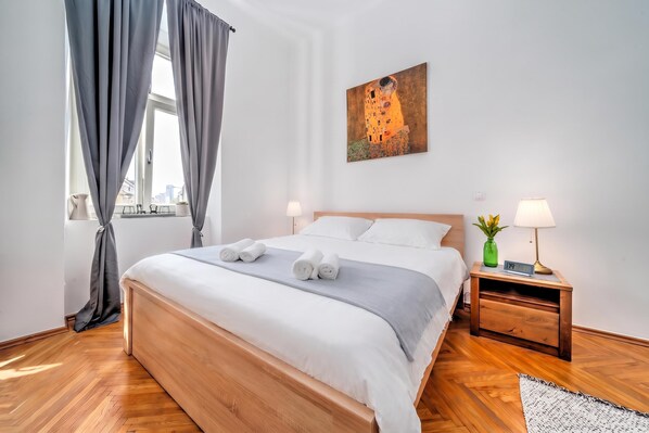 A hotel room with a large bed, two bedside tables, a painting on the wall, and a view of the city through the window.