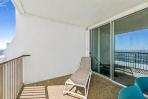 A balcony with a white chair, a blue chair, and a glass door leading to a terrace with a view of the sea.