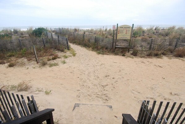 A sandy beach path leading to a dune crossing sign, surrounded by wooden fences and sparse vegetation.