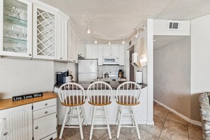 A compact kitchen with white cabinetry, a built-in microwave, and a coffee machine. There are three white chairs with wooden seats around a small table.